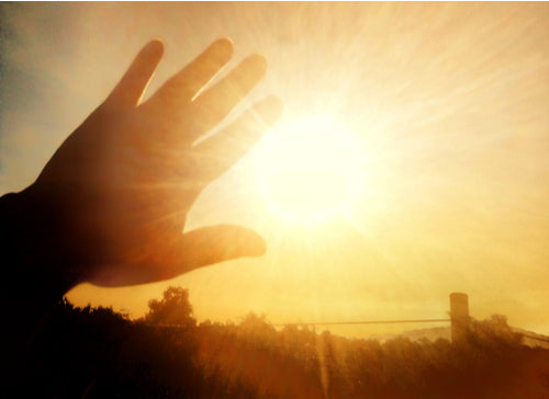 A silhouette of a hand reaching toward the sun outlined in warm light against a hazy horizon illustrating environment impact on skin.
