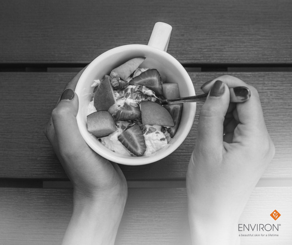 White ceramic mug filled with sliced fruit and yogurt held by hands with dark nail polish for winter energy boost