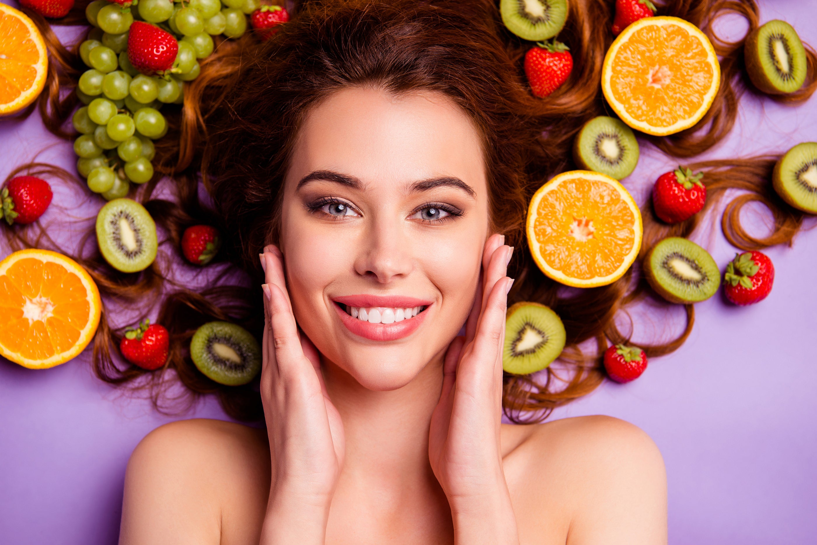 A woman with long wavy brown hair and radiant smile lies among vibrant fruits for a 2020 skin detox routine