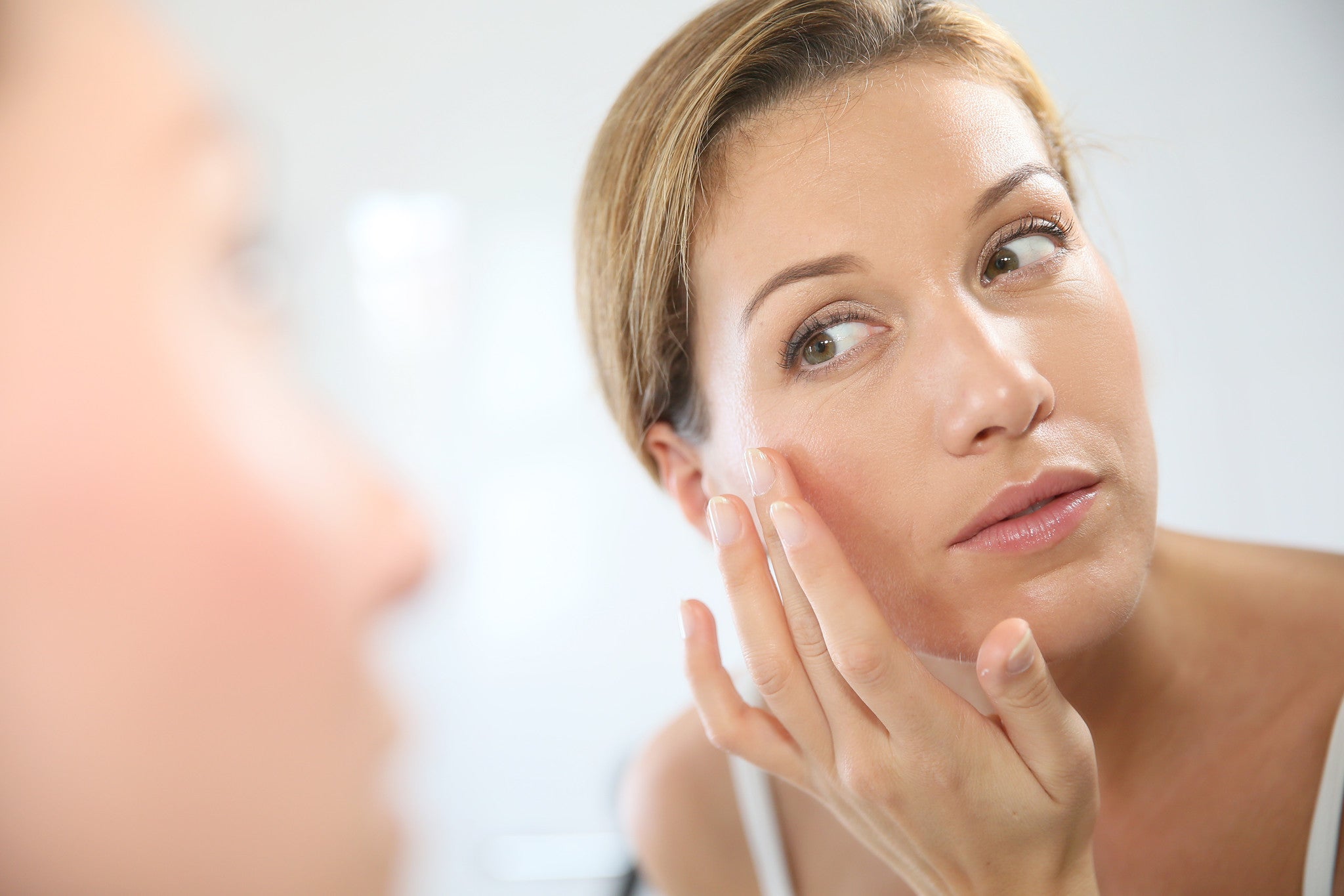 A woman with short blonde hair gently touches her face looking in the mirror for skin concern reflection.
