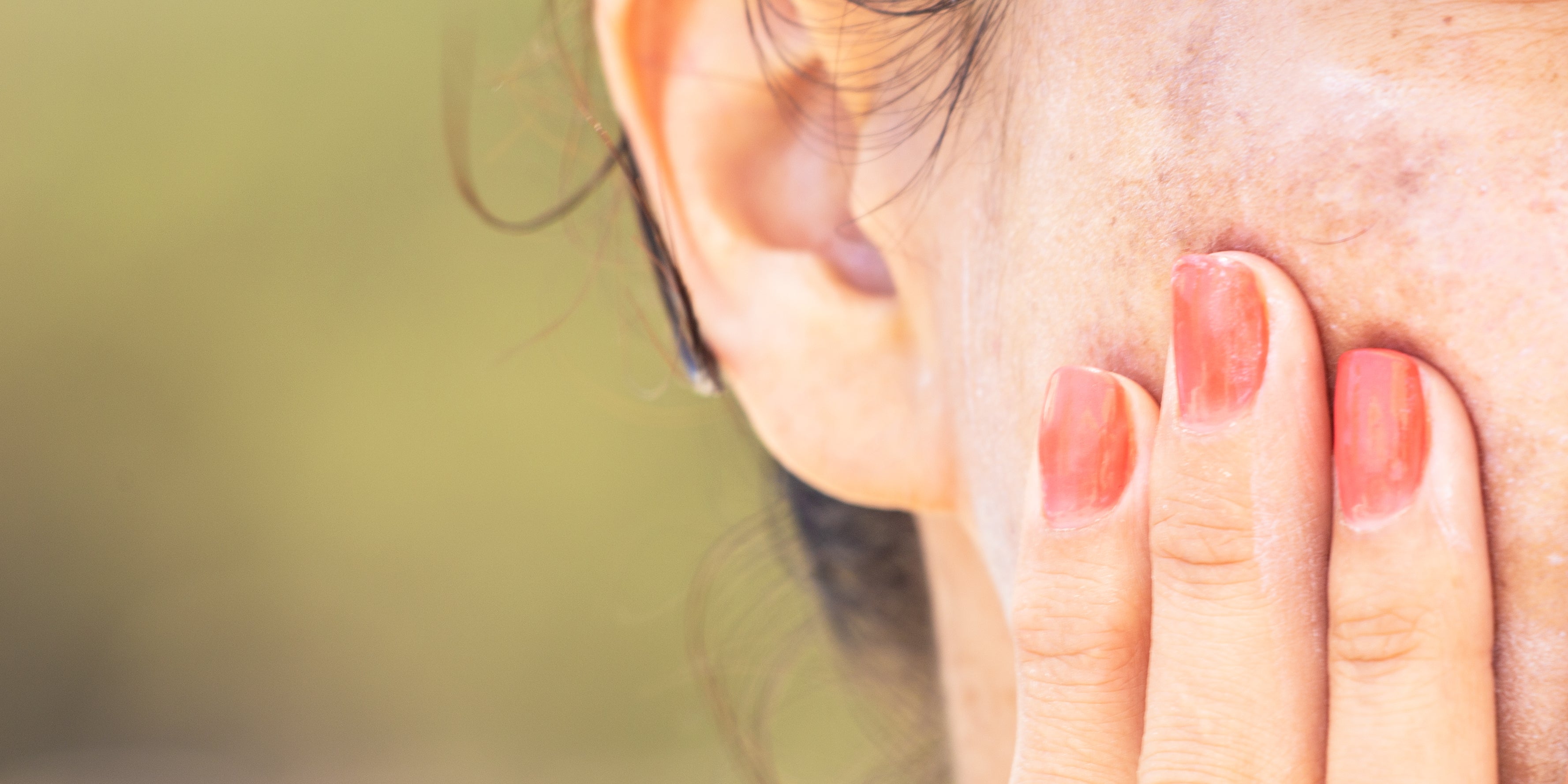 A hand with coral-painted nails gently touching skin near an ear for summer guide to even-looking complexion