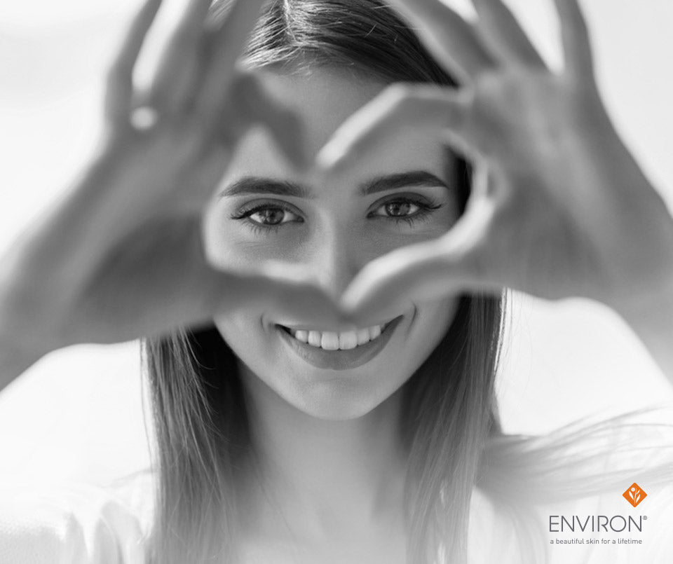 A woman with long hair smiling, hands forming a heart shape in front of her face, illustrating youthful-looking eyes.