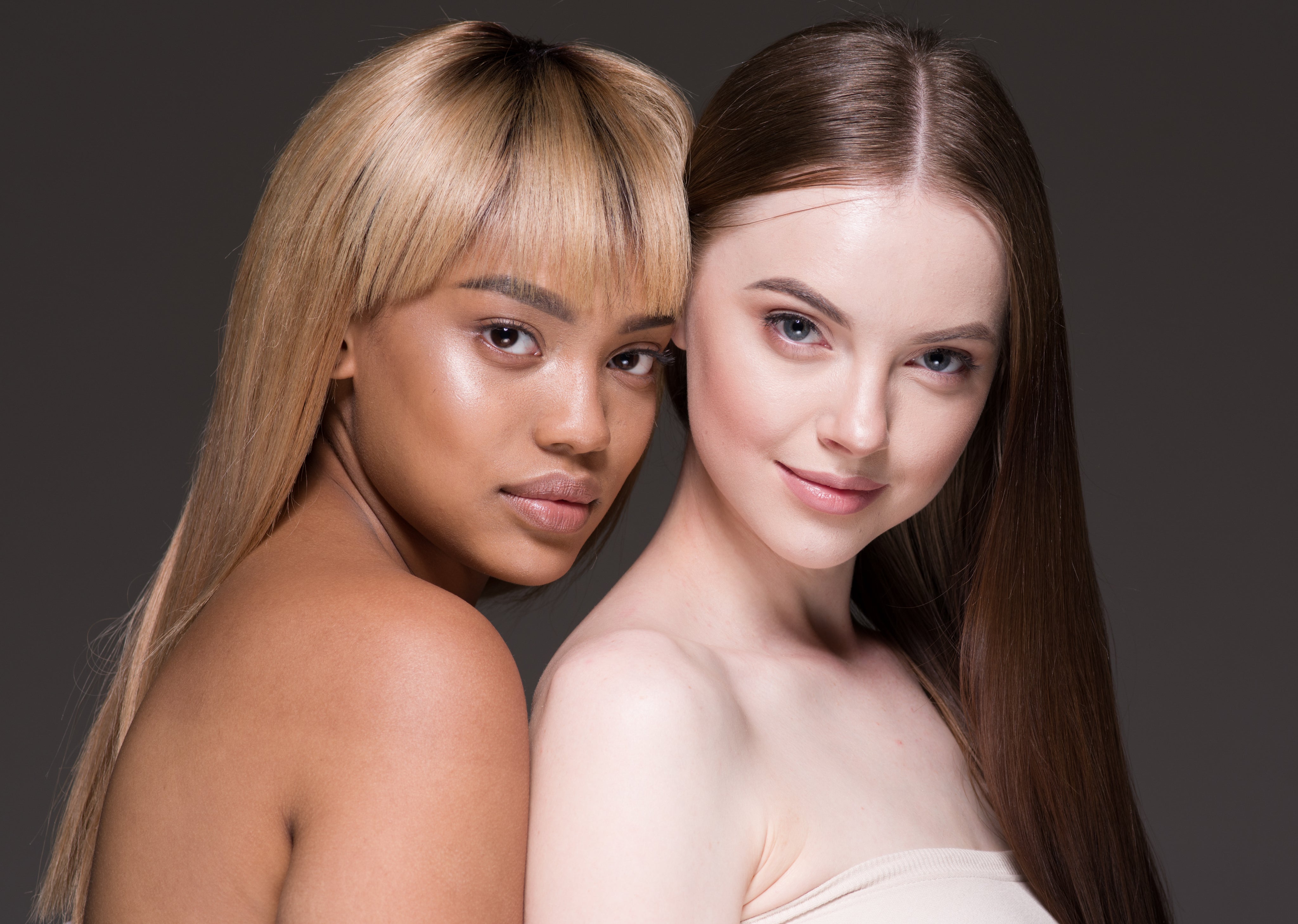 Two women with long, straight hair, one blonde with a blunt fringe and the other brunette, pose closely together against a neutral gray backdrop.
