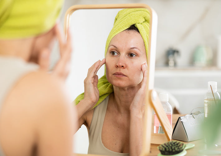 A woman with a bright lime green towel wrapped around her head looks in a wooden mirror touching her face in a skin care moment.