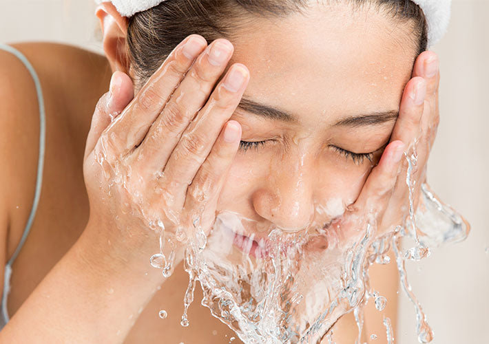 A woman wearing a white towel on her head washing her face with water in an article about suds and surfactants.