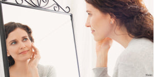 A woman with dark hair gazes at her reflection in a decorative black wrought iron mirror in an aging skin article.