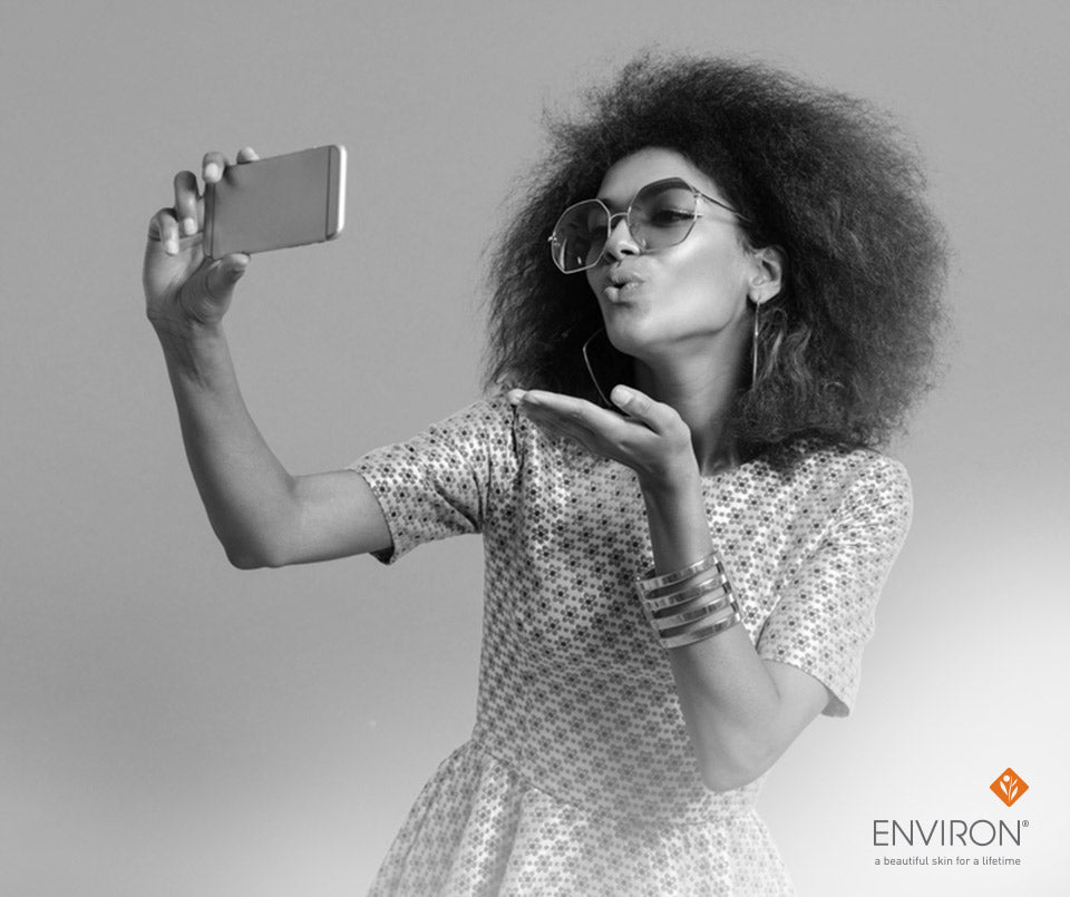 A woman with curly hair and sunglasses blowing a kiss while taking a selfie for love your skin article