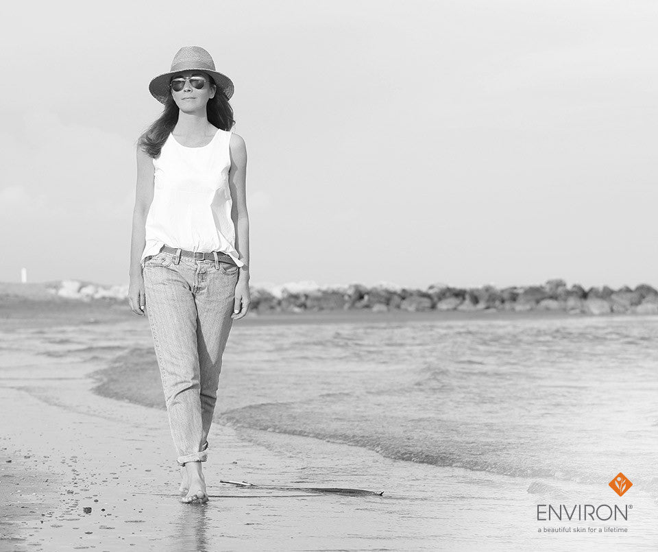 A woman wearing a straw hat and sunglasses walks barefoot along the shoreline promoting visibly luminous summer skin care