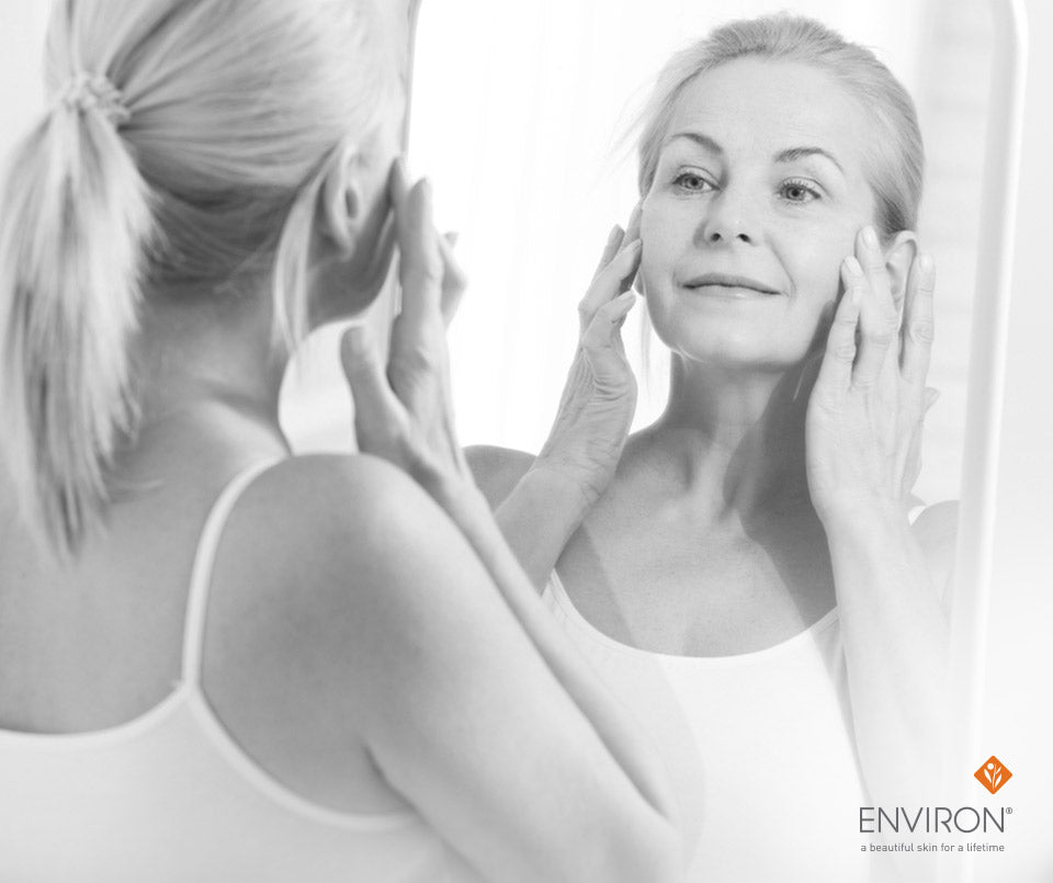 A woman with blonde hair in a ponytail wearing a white tank top gently touching her face while looking in the mirror
