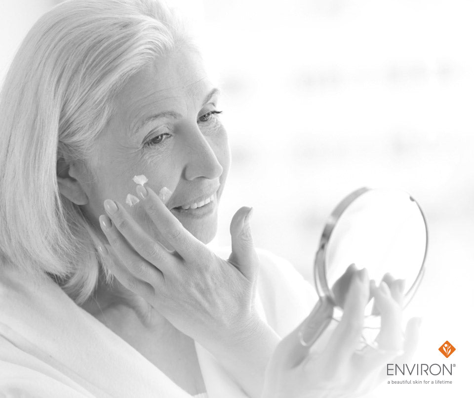 A woman with white hair gently applies soothing cream to her face while holding a small round mirror for sensitive skin.