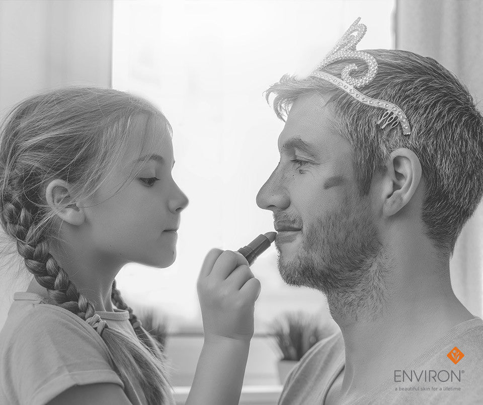 A man wearing a tiara has his lips painted by a young girl illustrating dads deserve a dewy glow too
