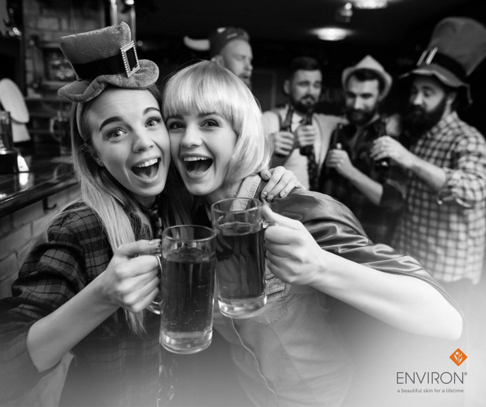 Two women in festive attire toast with tall clear glass mugs filled with dark liquid in a how to have great skin after a night out article