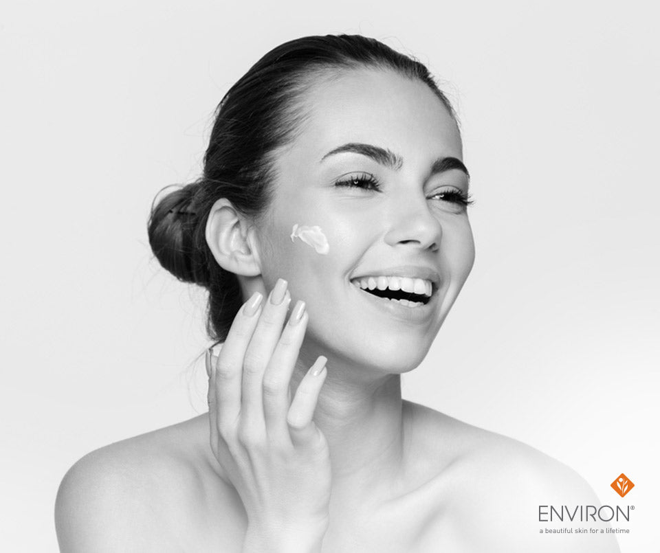 A woman with dark hair in a bun smiling while applying white cream to her cheek in wake up skin article.