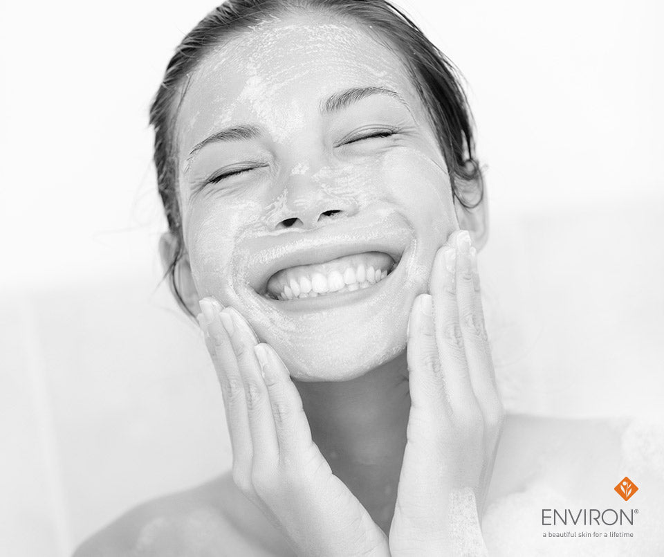 A woman smiling with eyes closed while applying a facial mask using interactive cleansing gel for skincare.