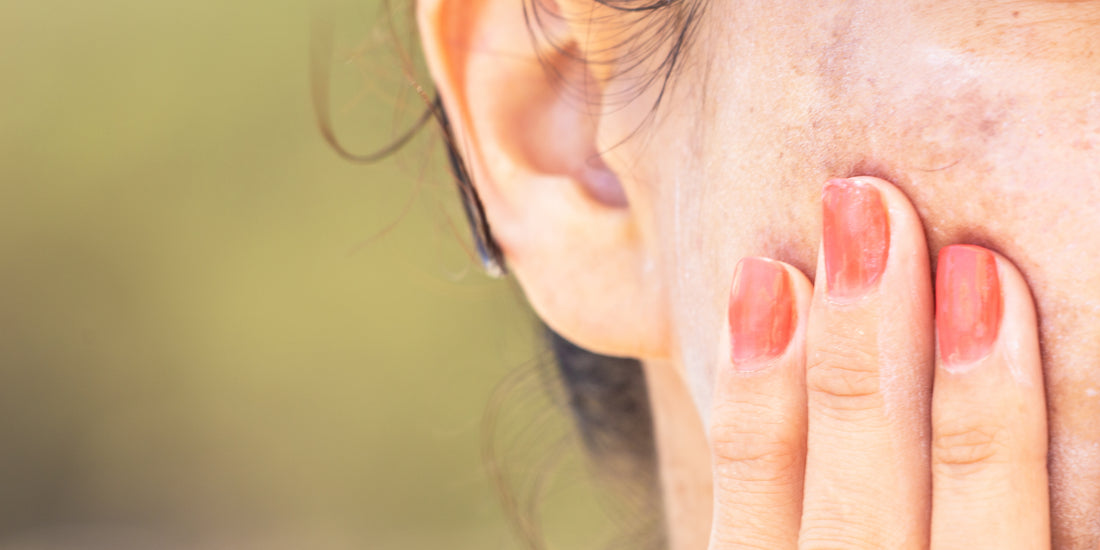 A hand with coral-painted nails gently touching skin near an ear for summer guide to even-looking complexion