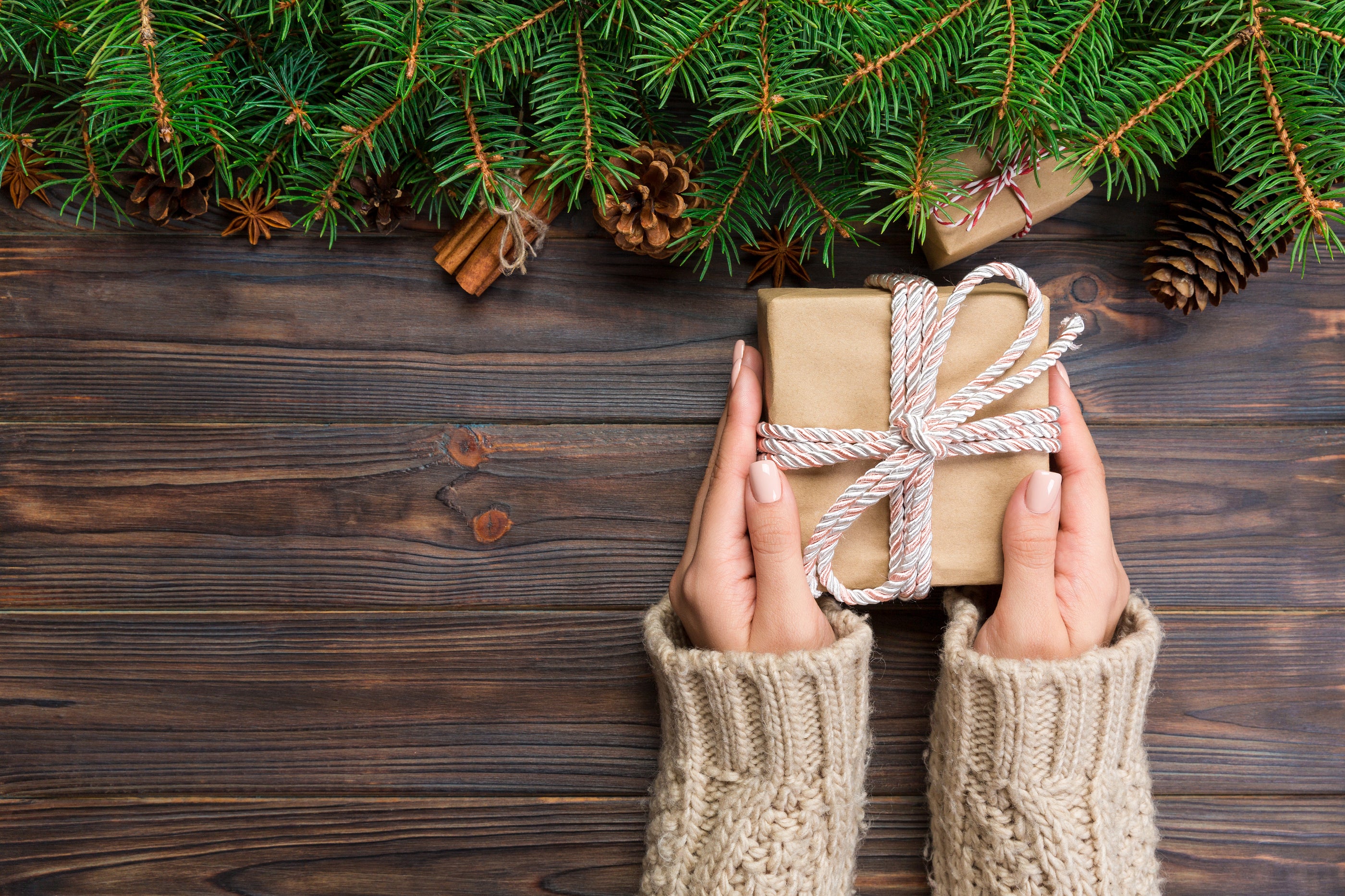 A beige knitted cable knit sweater worn by hands holding a brown paper wrapped gift with a white rope bow