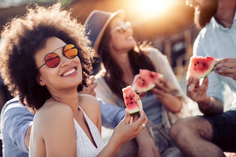 A woman with curly hair wearing round reflective orange sunglasses and a white knit top in a skin health diet article