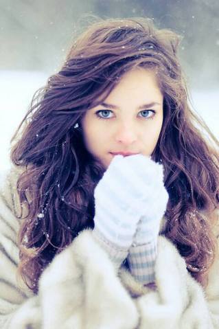 A woman with long wavy brown hair and blue eyes wears a fluffy beige coat and white knit gloves in the snow.