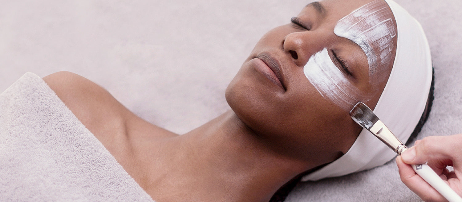 A white facial brush with a sleek, metallic handle is being used to apply a thick, creamy white mask.