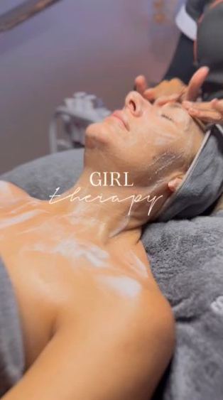 A woman reclines on a gray towel-covered treatment bed, her head wrapped in a dark gray towel with white text reading ’girl therapy’ across the forehead.