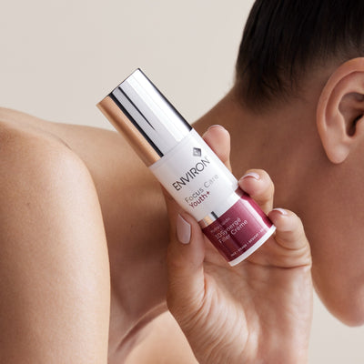 A sleek white and rose gold environ focus care youth+ 3d synergé filler crème bottle with a metallic cap, held gently in a hand.
