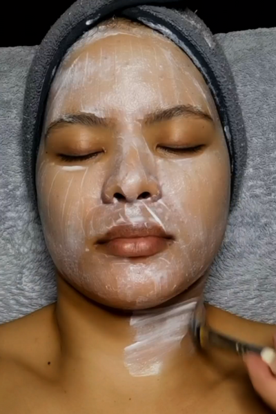 A person wearing a gray towel on their head while receiving a facial treatment with a white, translucent sheet mask being applied to their face.