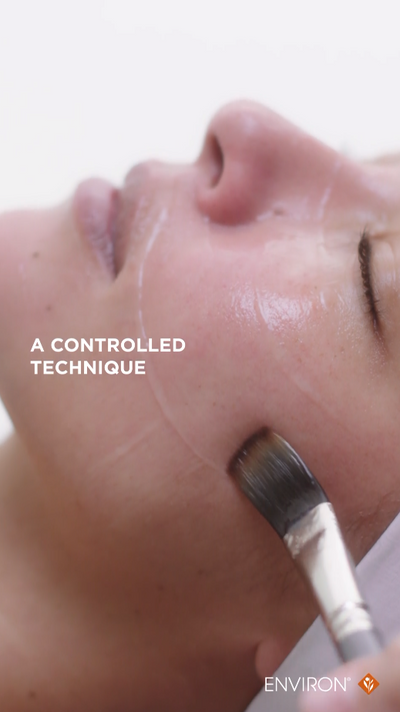 A sleek, silver-tipped facial brush with a black bristle head is being used to apply a glossy, translucent liquid treatment.