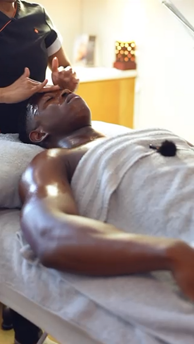 A man with dark skin lies on a massage table, covered by a white towel.