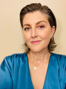A woman wears a sleek, satin blue blouse with ruched shoulders and a delicate gold circular pendant necklace.