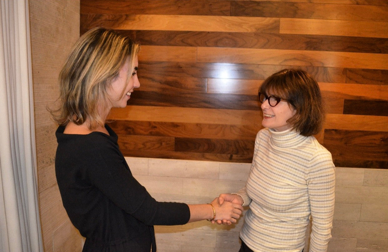 Two women are shaking hands in front of a wooden wall.
