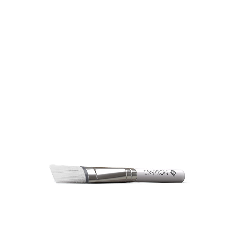 An Environ angled facial treatment brush with soft white bristles and a silver ferrule rests horizontally on a white background.