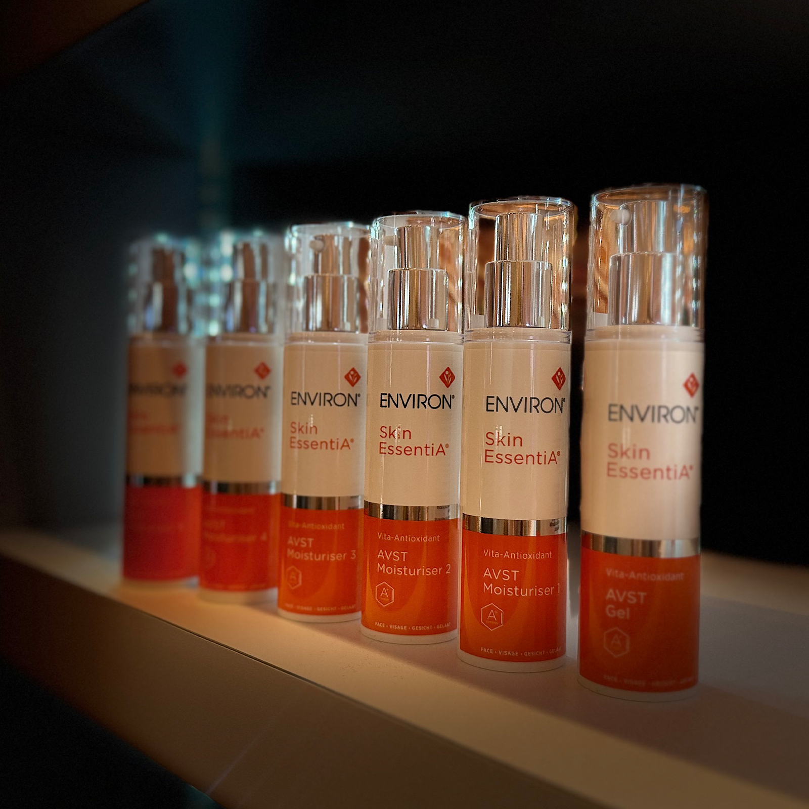 A row of environ skin essentia avst moisturiser bottles with clear glass bodies, silver pump tops, and labels featuring white, orange, and red branding.