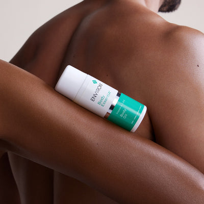 White and teal environment body essential vitamin a-care body oil bottle resting on a person’s arm.