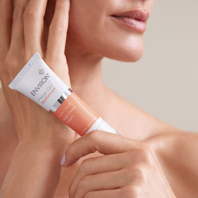A white and peach-colored environ focus care radiance+ multi-c action melts-even cream tube with a silver cap, held gently in hands.