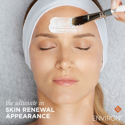 A white facial brush with a silver ferrule and black bristles is being used to apply a creamy product.