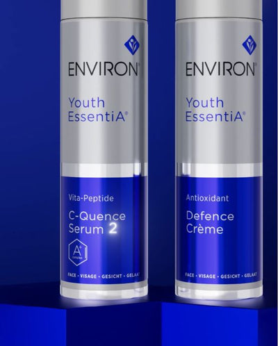 Two silver and blue environ youth essentia skincare bottles, one labeled ’c-quence serum 2’’ with vita-peptide and the other ’antioxidant defence crème,’ standing side by side on a reflective surface.