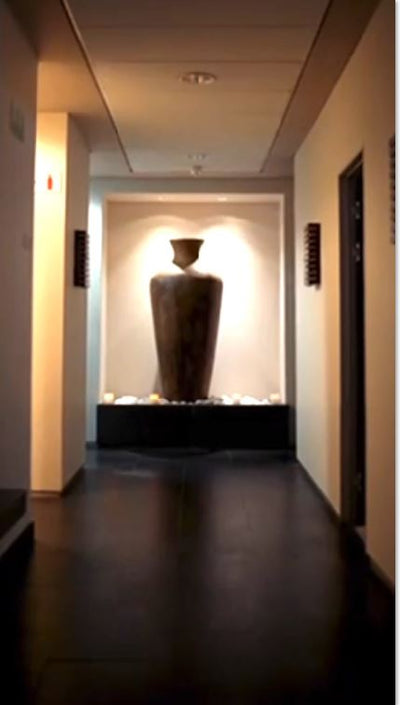 A tall, dark brown ceramic vase with a narrow neck and wide base sits prominently on a black pedestal in the center of a dimly lit hallway.