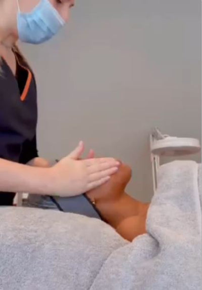 A person wearing a blue surgical mask and dark uniform with orange trim is gently massaging the leg of another individual who is lying down, covered by light gray fabric.