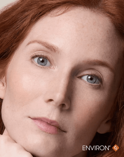 A close-up portrait of a woman with vibrant red hair and striking blue eyes, her face softly illuminated against a neutral background.