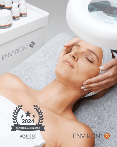A woman reclines on a treatment bed with her eyes closed, receiving an environ facial procedure under a white overhead light.