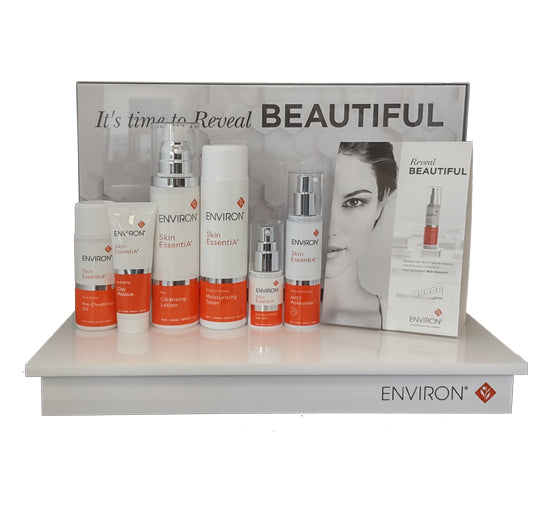 The Environ Skin EssentiA skincare collection, featuring various cleansers and moisturizers, stands neatly on a white retail display next to a "Reveal Beautiful" promotional brochure.